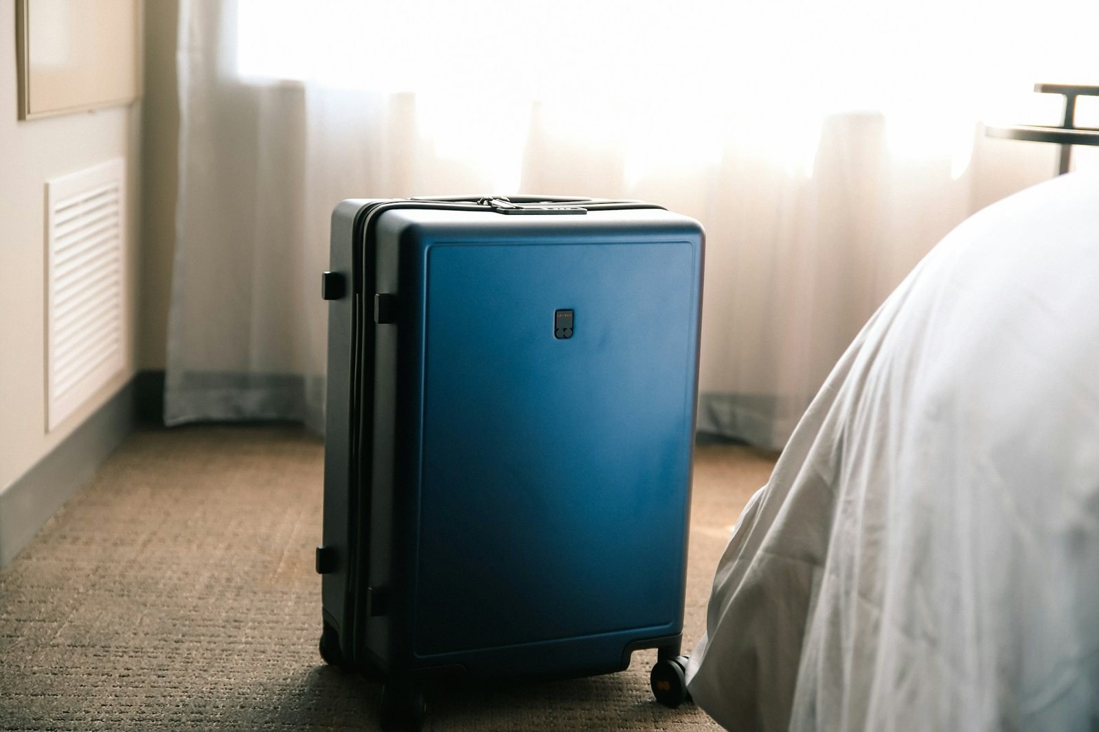 Blue hard-shell suitcase standing beside a hotel bed