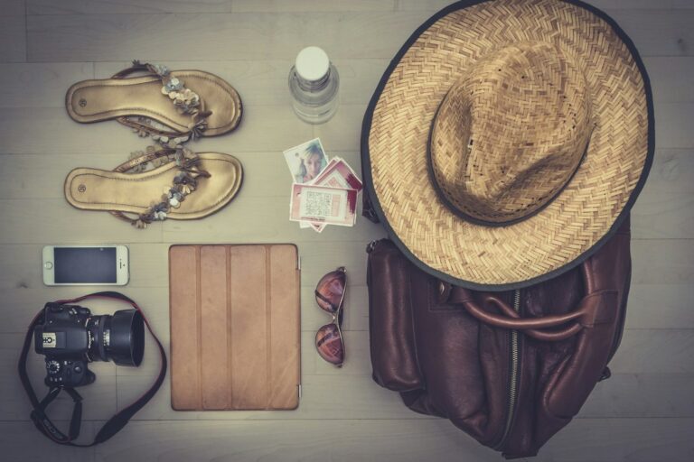 Flat lay of travel accessories including a hat, sandals, phone, tablet, camera and bag