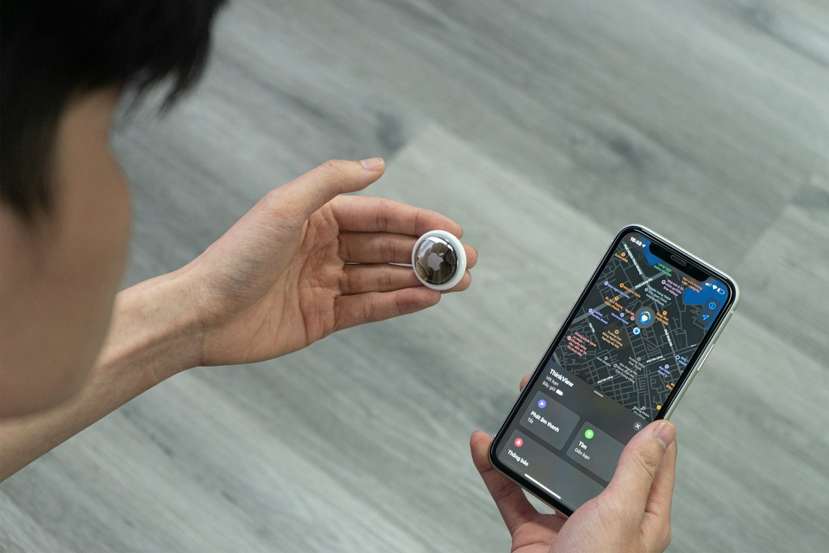 Hand holding a tracker tag next to a phone showing a map