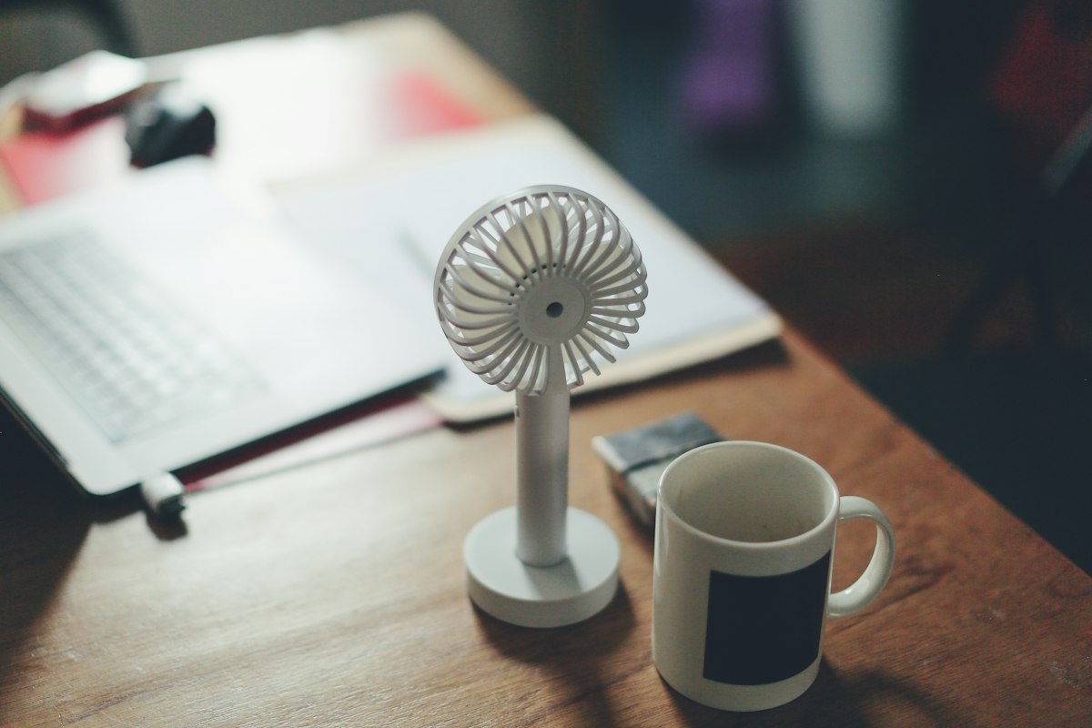 Small portable fan on a desk next to a laptop and mug