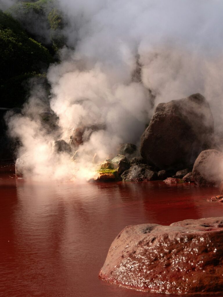 Steam billowing over red-tinted hot spring water and rocks at the Hells of Beppu