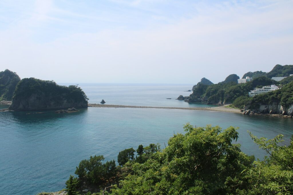 A narrow sandbar linking a rocky wooded islet to the shore in a blue bay on the Izu coast